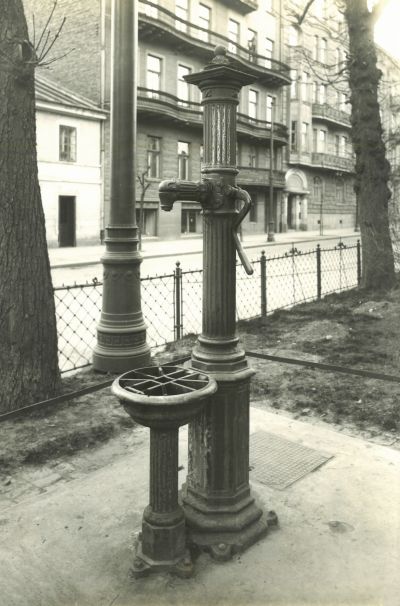 Studnia publiczna. Hydrant z pompą zlokalizowany przy budynkach mieszkalnych. Zdjęcie czarnobiałe.