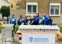 Wolonatriusze w pelerynach stoją przed stołem, na którym znajdują się kubeczki z wodą. Z boku stoi też nalewak do wody.
