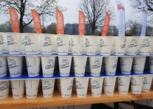 Kubeczki z wodą umieszczone są na stole w trzech rzędach, jeden na drugim. W tle widać powiewające flagi.