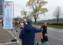 Biegnący maratończyk dostaje od wolontariusza kubek z wodą. Z boku stoi flaga, a w tle widać biegnących zawodników. 