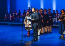 Na scenie stoi Piotr Ziętara i przemawia do mikrofonu w tle ukraiński zespół pieśni i tańca.